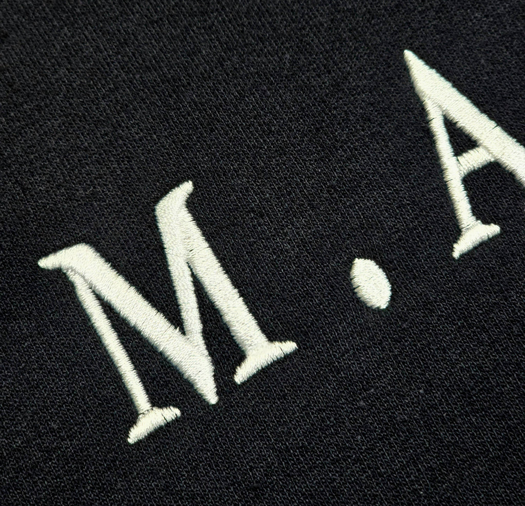 Macro close-up of white embroidered M.A.N.F.Y. lettering on black premium fabric