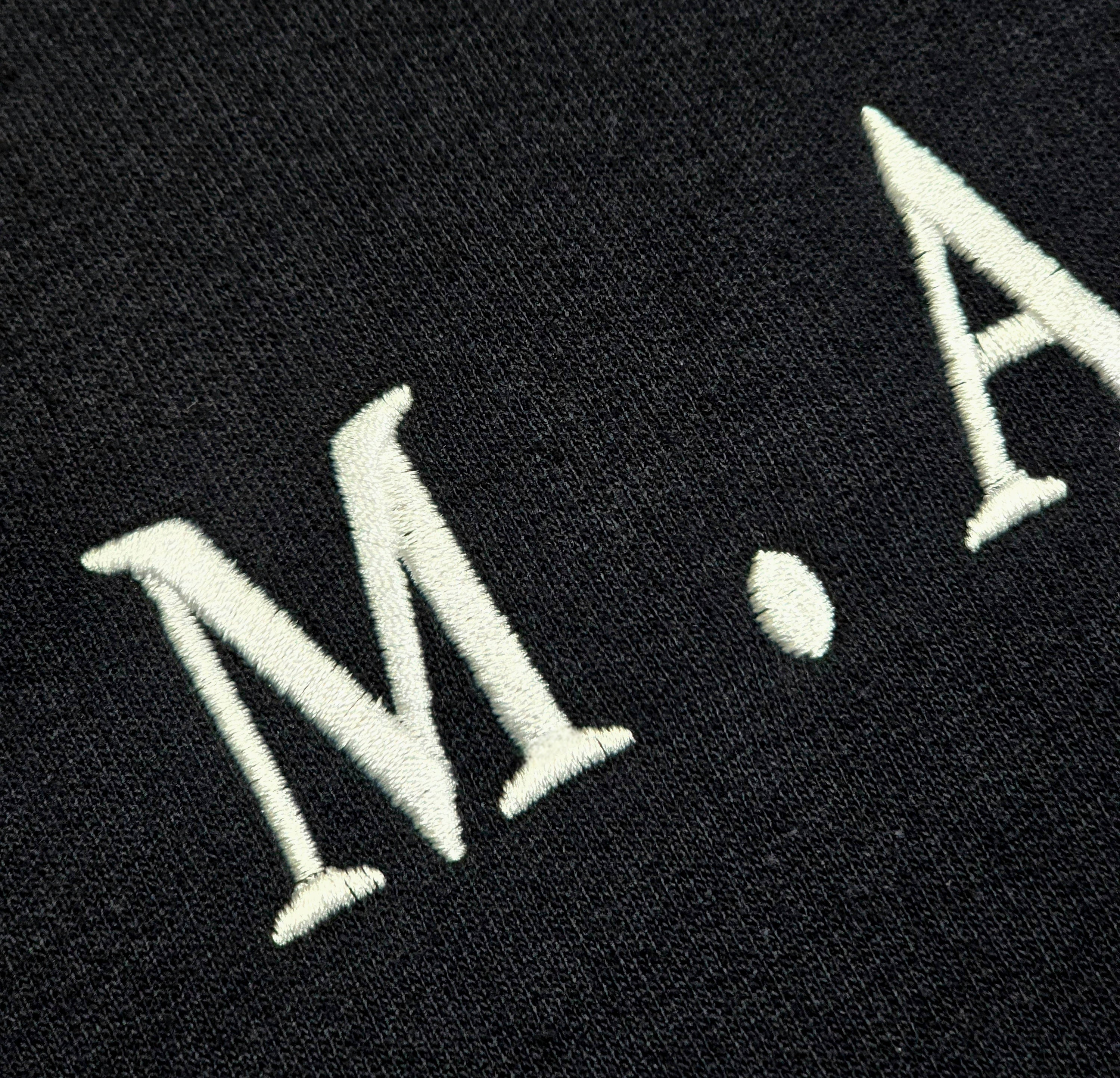 Macro close-up of white embroidered M.A.N.F.Y. lettering on black premium fabric